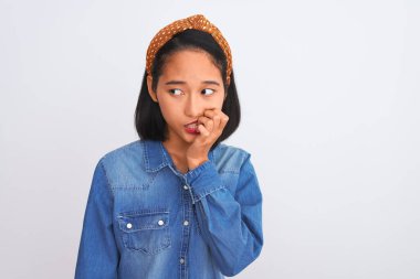 İzole edilmiş beyaz arka planda duran kot gömlek giyen genç ve güzel Çinli kadın elleri ağız yiyen tırnaklarla stresli ve gergin görünüyor. Endişe sorunu.