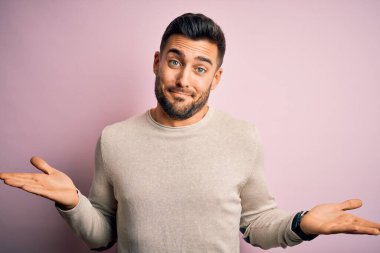 Young handsome man wearing casual sweater standing over isolated pink background clueless and confused expression with arms and hands raised. Doubt concept. clipart