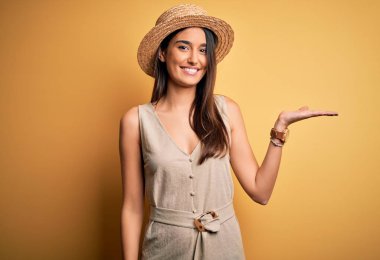 Genç, güzel, esmer bir kadın tatilde. Günlük elbiseler ve şapkalar takıyor. Neşeli bir şekilde gülümsüyor ve avuç içi kameraya bakıyor..