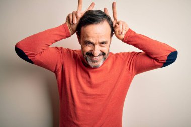 Orta yaşlı, turuncu süveter giyen, izole edilmiş beyaz arka planda duran, parmakları kafasında tavşan kulağı olan, neşeli gülümseyen, komik ve çılgın bir adam.