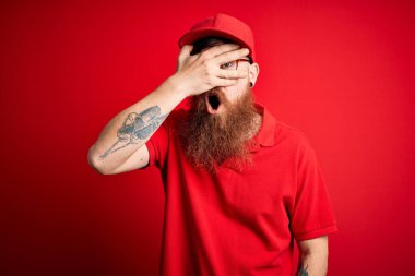 Gözlüklü ve kırmızı şapkalı genç teslimatçı, izole edilmiş arka planda, şokla yüzünü ve gözlerini kapayarak utangaç ifadesiyle parmaklarının arasından bakıyor..