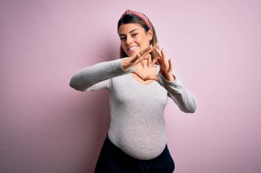 Genç, güzel, esmer bir kadın hamile, pembe arka planda bebek bekliyor. Aşkla gülümsüyor, elleriyle kalp sembolü yapıyor. Romantik konsept.