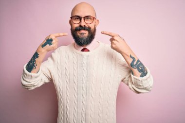 Sakallı, dövmeli, pembe arka planda gözlüklü ve süveterli neşeli gülümseyen ve parmaklarını dişleriyle ve ağzıyla işaret eden yakışıklı kel adam. Diş sağlığı konsepti.
