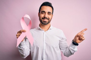 Sakallı, pembe kanser kurdelesini soyutlanmış arka planda tutan genç ve yakışıklı bir adam. Elleri ve parmakları kenarı işaret ederek çok mutlu.