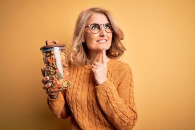 Orta yaşlı güzel sarışın kadın sarı arka planda İtalyan makarnası kavanozu tutuyor. Ciddi bir yüz. Soru sormayı düşünüyor.