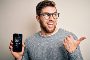 Sarışın, sakallı, mavi gözlü, gözlüklü, kırık akıllı telefon tutan ve gülümseyen bir yüzü olan genç bir adam.