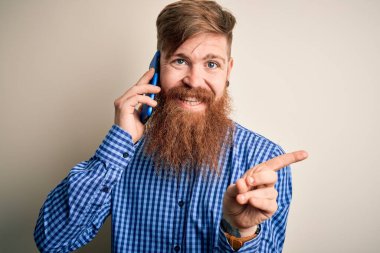 Kızıl saçlı, sakallı bir İrlandalı adam, izole bir arka planda akıllı telefondan konuşuyor. Elle ve parmaklarıyla işaret ederek çok mutlu.