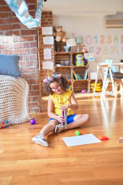 Gözlüklü ve tek boynuzlu at diademi takmış güzel bir çocuk. Anaokulunda kağıt ve kalem kullanarak yerde oturuyor.