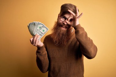 Kızıl saçlı, sakallı, sarı arka planda 1 dolarlık banknotlar tutan, gülümseyen, el üstünde el işareti yapan, parmaklarının arasından bakan İrlandalı bir adam.
