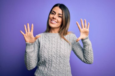 İzole edilmiş mor arka plan üzerinde duran sıradan bir süveter giyen genç ve güzel bir kadın on numaralı parmaklarıyla işaret ederken kendinden emin ve mutlu bir şekilde gülümsüyor..
