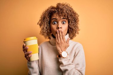 Genç Afro-Amerikalı kıvırcık kadın sarı arka planda bir fincan kahve içiyor. Ağzı kapalı, utancından ağzı açık, korku ifadesi, sessizlikten korkmak, gizli bir konsept.