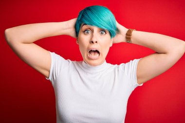 Mavi saçlı genç bir kadın kırmızı arka planda sıradan tişörtler giyiyor. Elleri kafasında çılgın, korkmuş ve ağzı açık şoktan korkan bir kadın.