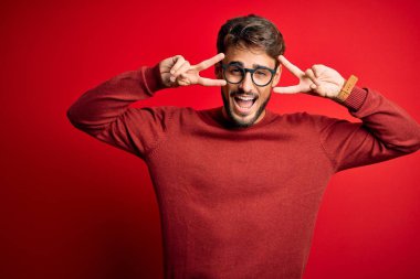 Sakallı, gözlüklü ve süveterli genç adam kırmızı arka planda duruyor. Barış sembolü yapıyor. Yüzünde parmaklar, gülümseyerek zaferini gösteriyor.
