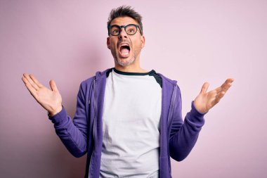 Pembe arka planda duran mor eşofman üstü ve gözlüklü genç yakışıklı adam deli deli deli bağırıyor ve agresif bir ifade ve kollar havada bağırıyor. Hayal kırıklığı konsepti.