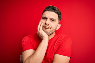 Mavi gözlü genç adam kırmızı izole edilmiş arka plan üzerine günlük tişört giyiyor. Kollarını kavuşturarak depresyondan sıkılmış ve yorgun görünüyor..