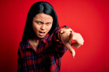 Genç, güzel, Çinli bir kadın, günlük gömleğini kırmızı arka planda izole edilmiş bir şekilde giyiyor. Kameraya kızgın ve sinirli bir şekilde bakıyor.