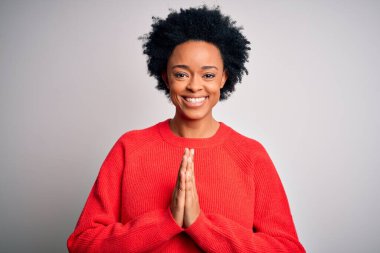 Kıvırcık saçlı, kırmızı kazak giyen, elleri birarada dua eden ve kendinden emin bir şekilde gülümseyen Afro-Amerikan bir kadın..