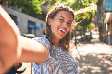 Genç, güzel bir kadın mutlu bir şekilde gülümsüyor. Güneşli bir yaz gününde şehrin sokaklarında yürüyor. Akıllı telefon kullanarak selfie çekiyor.