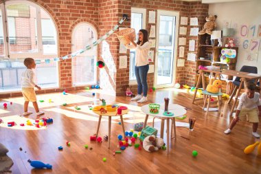 Anaokulunda bir sürü oyuncağın etrafında top ve hasır sepet kullanarak basketbol oynayan güzel öğretmenler ve yeni yürümeye başlayan çocuklar.