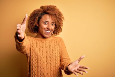 Kıvırcık saçlı, sıradan bir kazak giyen, kollarını açıp kameraya gülümseyen güzel Afro-Amerikan kadın. Mutluluğu kucaklayan neşeli bir ifade..