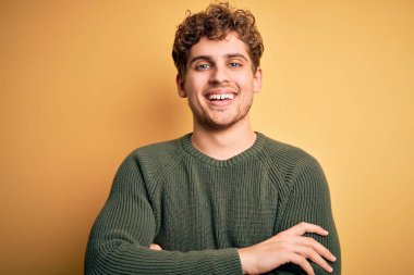 Sarışın, kıvırcık saçlı, sarı arka planda yeşil kazak giyen, kollarını kavuşturup kameraya bakan genç, yakışıklı bir adam. Pozitif kişi.