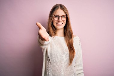 Genç, güzel, kızıl saçlı bir kadın pembe arka planda günlük süveter ve gözlük takıyor. Gülümsüyor, gülümsüyor, avuç içi kadar yardım ve kabul görüyor..