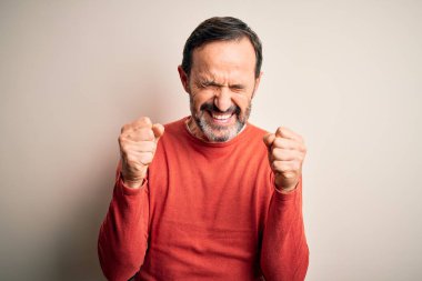 Orta yaşlı, turuncu süveter giyen, izole edilmiş beyaz arka plan üzerinde duran, kolları havada ve gözleri kapalı zaferi kutlayan, mutlu bir adam. Kazanan konsept.