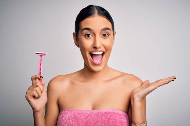 Young beautiful brunette woman wearing towel using razor for depilation very happy and excited, winner expression celebrating victory screaming with big smile and raised hands clipart