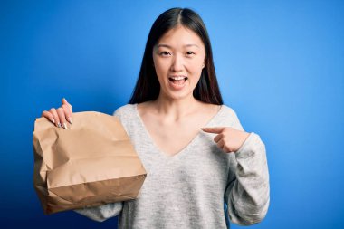 Asyalı genç bir kadın, servis paketi taşıyor. Paket yemek için mavi izole edilmiş arka planda. Şaşırmış yüz ifadesi kendini gösteriyor.