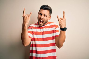 Sıradan çizgili tişört giyen yakışıklı genç adam izole edilmiş beyaz arka plan üzerinde duruyor çılgın bir ifadeyle elleri havada rock sembolü yapıyor diye bağırıyor. Müzik yıldızı. Ağır müzik konsepti.