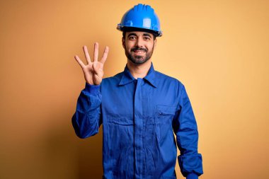 Sakallı tamirci mavi üniformalı ve kasklı sarı arka planda kendini beğenmiş ve mutlu bir şekilde gülümserken parmaklarını dört numarayı gösteriyor..