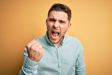 Mavi gözlü genç bir iş adamı sarı arka planda zarif yeşil tişörtler giyiyor kızgın ve kızgın. Öfkeyle bağırırken yumruğunu kaldırıyor. Öfke ve agresif kavram.