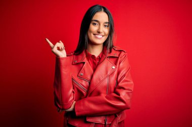 Genç, güzel, esmer, günlük ceket giyen, kırmızı arka planda duran, yüzünde kocaman bir gülümseme olan, elleri ve parmakları kameraya bakan bir kadın..