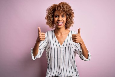 Kıvırcık saçlı güzel Afro-Amerikalı kadın pembe arka plan başarısı tabelasının üzerine çizgili tişört giyiyor. Elleriyle olumlu jest yapıyor, gülümsüyor ve mutlu. Neşeli ifade ve kazanan jest.