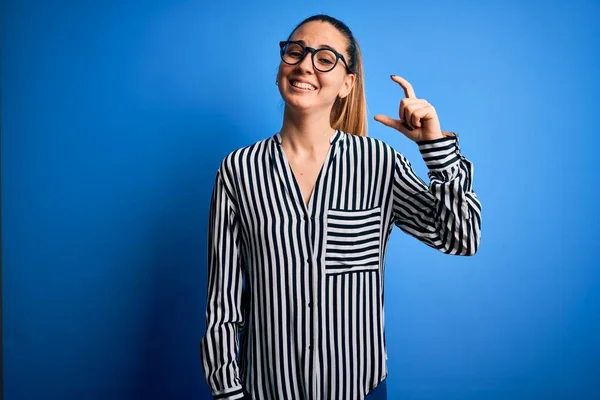 Beautiful blonde woman with blue eyes wearing striped shirt and glasses over blue background smiling and confident gesturing with hand doing small size sign with fingers looking and the camera. Measure concept.