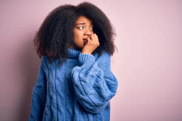 ピンクの背景に冬のセーターを着てアフロの髪を持つ若い美しいアフリカ系アメリカ人女性は 口の中に爪をかむ上で手でストレスと緊張しています 不安の問題 — ストック写真