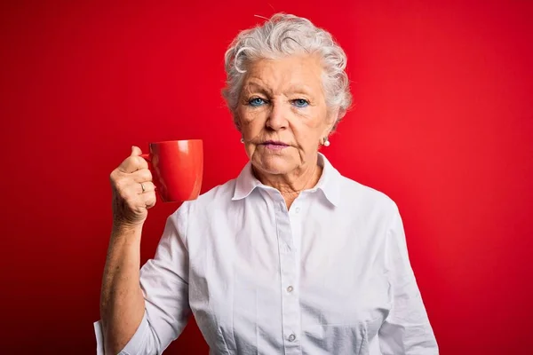年长而漂亮的女人喝着一大杯咖啡 站在孤立的红色背景上 脸上带着自信的表情 满不在乎地严肃思考着 — 图库照片
