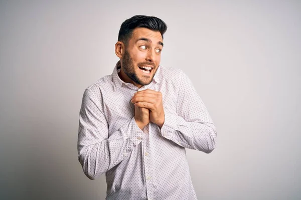 Young handsome man wearing elegant shirt standing over isolated white background laughing nervous and excited with hands on chin looking to the side