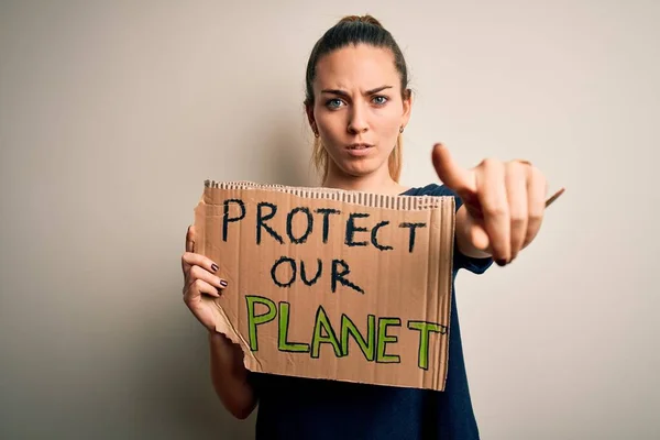 Young beautiful blonde woman with blue eyes asking for protect planet holding banner pointing with finger to the camera and to you, hand sign, positive and confident gesture from the front