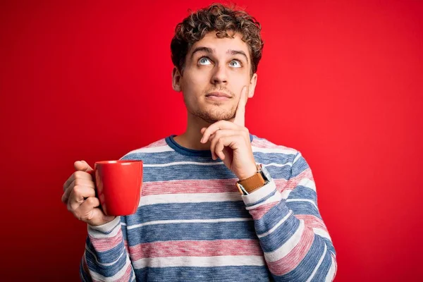 Young blond man with curly hair drinking cup of coffee standing over red background serious face thinking about question, very confused idea