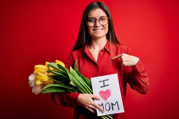 Young beautiful woman with blue eyes holding love mom message and tulips on mothers day with surprise face pointing finger to himself