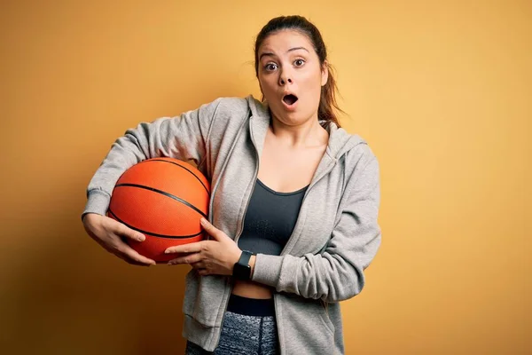 若い美しいブルネットのスポーツウーマンは 黄色の背景の上にバスケットボールボールを持っている驚きの顔でショックで怖がって 恐れと恐怖の表現で興奮 — ストック写真