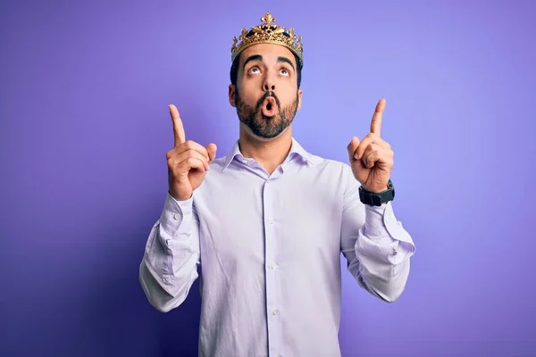 年轻英俊的大胡子男子 头戴金冠 头戴紫色背景的王冠 惊讶地抬起头 用手指和胳膊指指着 — 图库照片