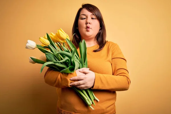 Beautiful plus size woman holding romantic bouquet of natural tulips flowers over yellow background making fish face with lips, crazy and comical gesture. Funny expression.