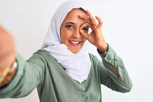 若いですアラブ女性身に着けていますHijab取ります自画撮り写真上の隔離された背景とともに幸せな顔笑顔行いますOk Signとともに手で目を通して指 — ストック写真