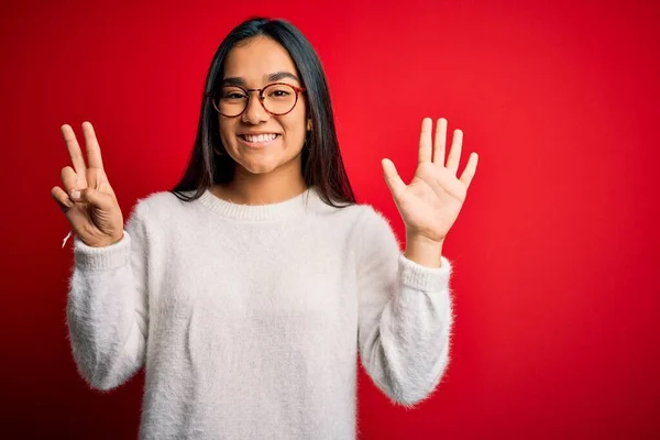 若いです美しいですアジアの女性身に着けていますカジュアルセーターと眼鏡上の赤い背景表示と指差しますと指番号7とともに笑顔自信と幸せ — ストック写真