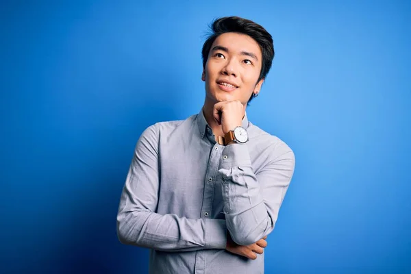 若いハンサムな中国人男性の質問について考えてあごの手で隔離された青の背景の上に立ってカジュアルなシャツを着て 集中的な表現 笑顔と思慮深い顔 疑いの概念 — ストック写真