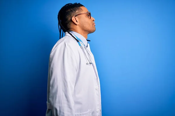 Young african american doctor man with dreadlocks wearing stethoscope and glasses looking to side, relax profile pose with natural face with confident smile.