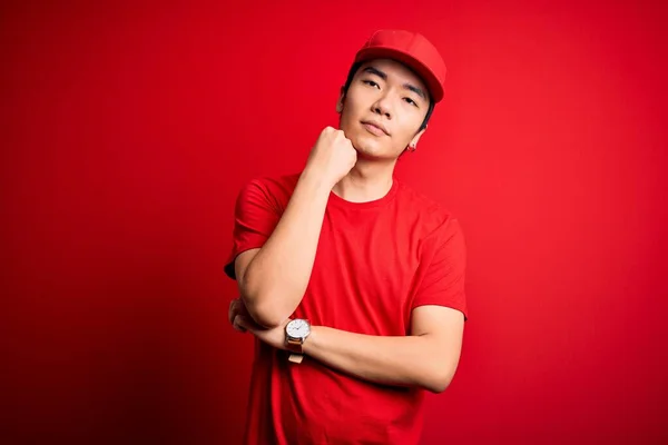 Young handsome chinese delivery man wearing cap standing over isolated red background with hand on chin thinking about question, pensive expression. Smiling with thoughtful face. Doubt concept.