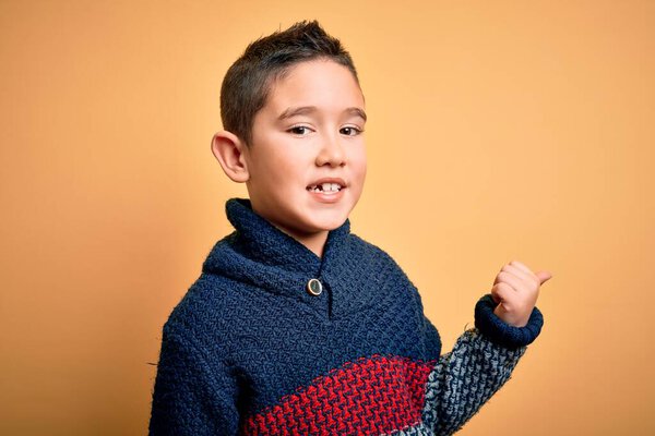 Маленький мальчик в зимнем свитере на желтом изолированном фоне улыбается со счастливым лицом, глядя и указывая на сторону с большим пальцем вверх
.